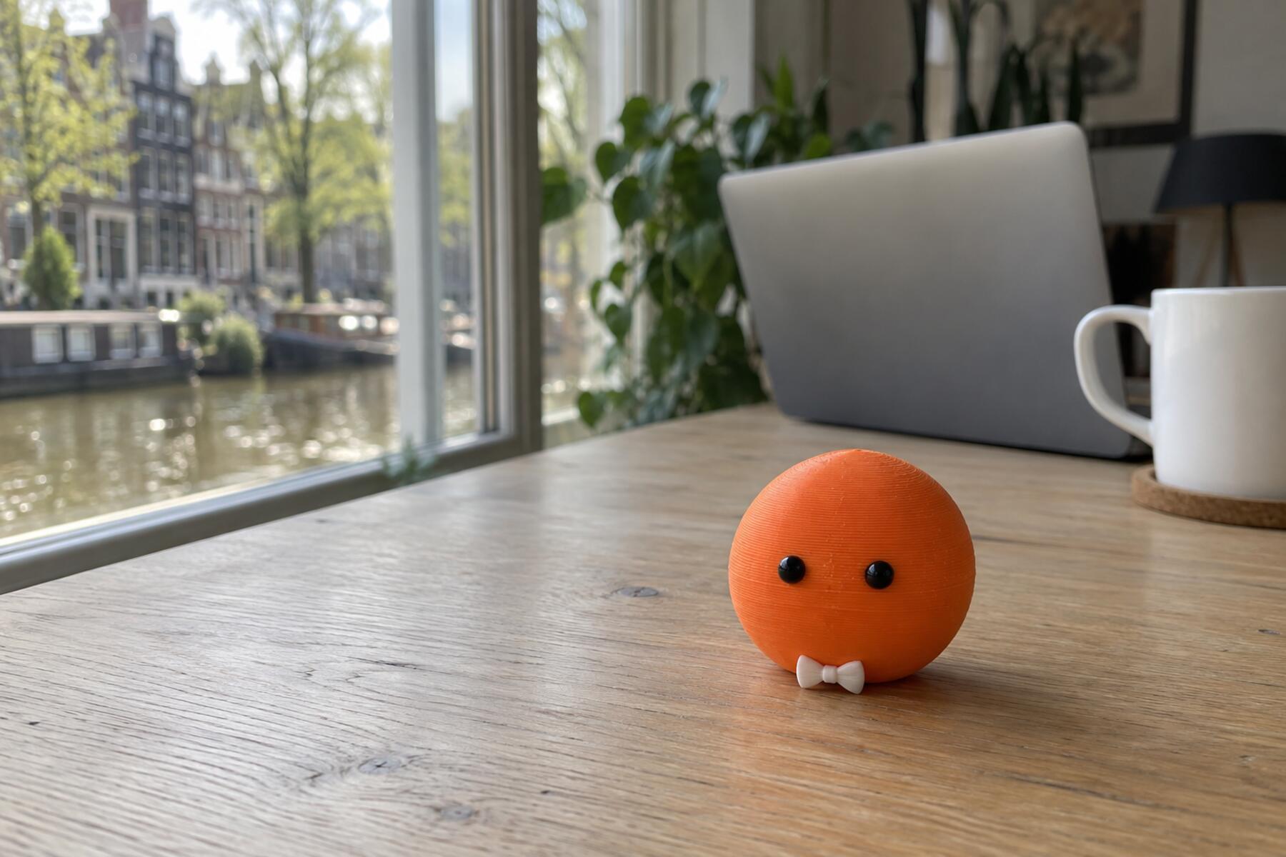 Een klein oranje figuurtje met twee oogjes en een wit strikje op een houten bureau, met daarachter een laptop en de gracht.